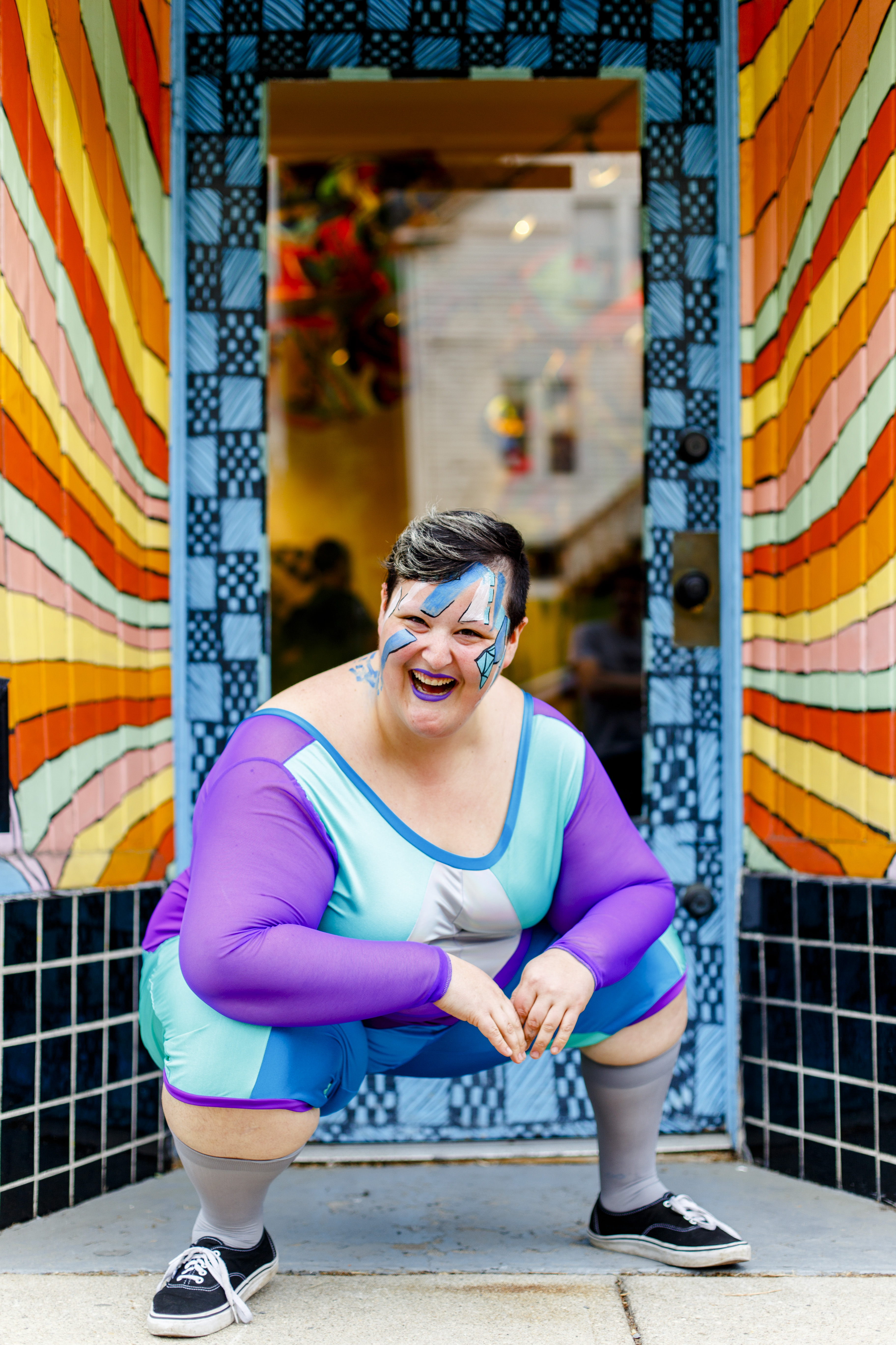 Sea Sprang squats down as they laugh in front of a doorway wearing a purple and blue geometric patterned piece with high socks and black sneakers. Photo credit Grace DuVal.