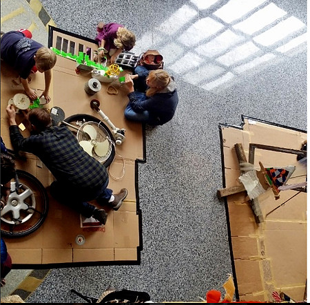 An image of for people working on the ground on flattened boxes. They are surrounded by miscellaneous items such as tires, a fan, and bright green tape.