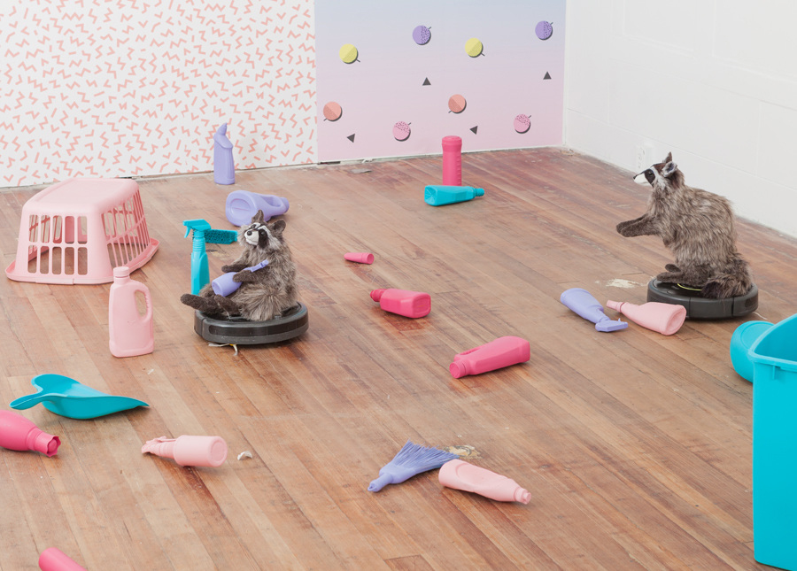 Two of the raccoons on Roombas moving in front of a wall with a pink chevron pattern. One is reclined and holds one of the bottles, the other is standing with its arms outstretched. Lux, Maria. “Dominus.” 2017. installation view. “Dominus,” DEMO Project, Springfield, IL. Courtesy of artist. Photos: Will Arnold.