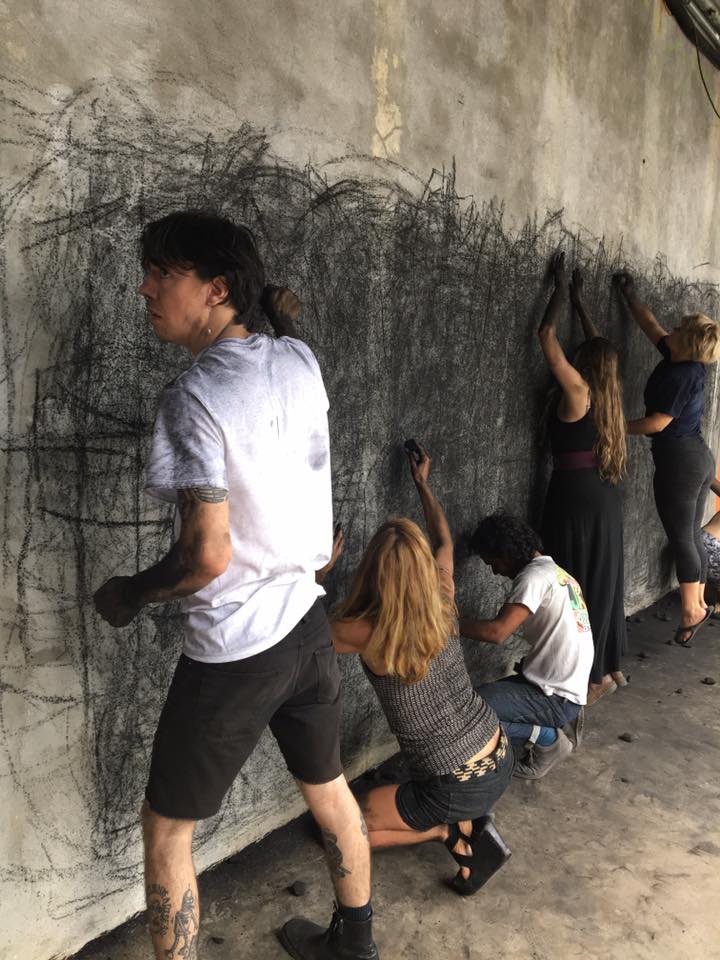 An image of five people in front of a wall, using wide gestures and positions to draw with charcoal in an abstract manner.