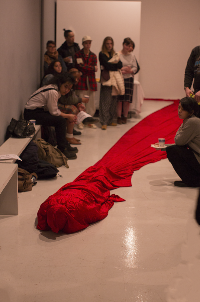 A red cloth is draped around the artist as they lie on the floor—the cloth is trailing behind them as a crowd of onlookers fill up the remainder of the gallery space. Photo by SAIC Documentation Team 