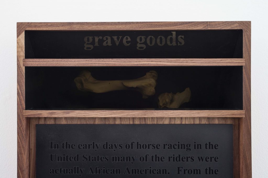 Three stacked horizontal panels, black and framed with brown wood; with text at the top reading "Grave Goods". Under that is a panel with an image of two horse bones, then finally a large panel with a long paragraph of text in black on a black background.