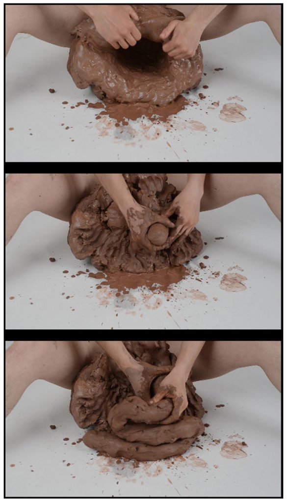 Courtesy of the artist. A still from the "Mature Female" series features the artists legs spread open with a large mound of clay in the center. Two hands are shaping the clay into a phallic shape. 