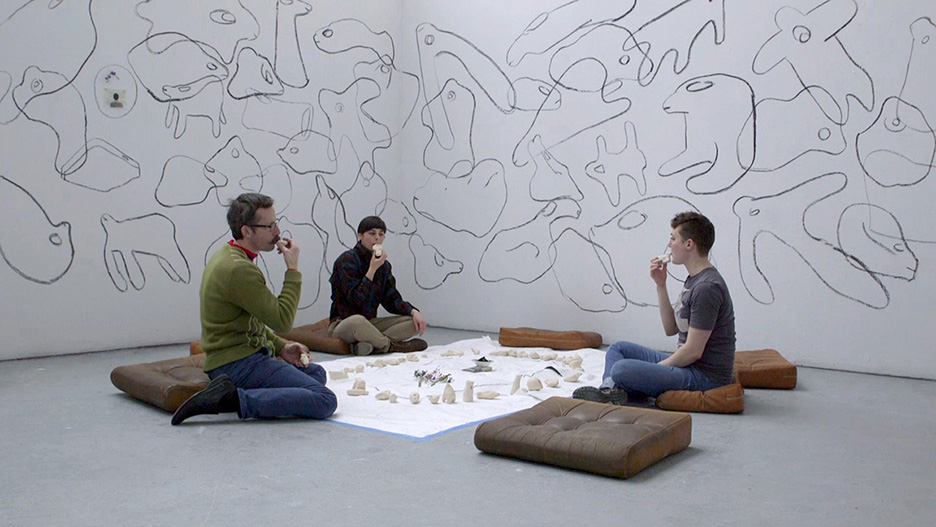 Courtesy of the artist. Three figures sit in the middle of a room in front of several clay whistles. They are each holding one to their mouth. 