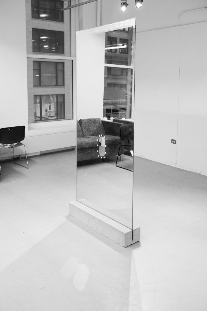 Courtesy of the artist. A long, tall mirror sits in the middle of a gallery space with a hole through the center. 