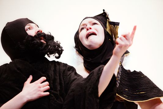 This is a color photograph still from the performance "Play." Two figures, draped in black clothing are shown. The one on the left is wearing a fake black beard and pointing with their left hand. The one on the right has a black head covering and appears to be wailing in despair.