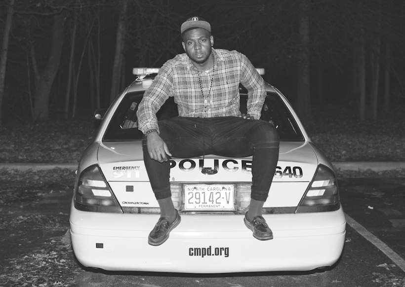 Courtesy of the artist. The back of a cop car faces the camera where the artist is seated on the back. His legs are open and he is wearing a long flannel shirt and hat. 