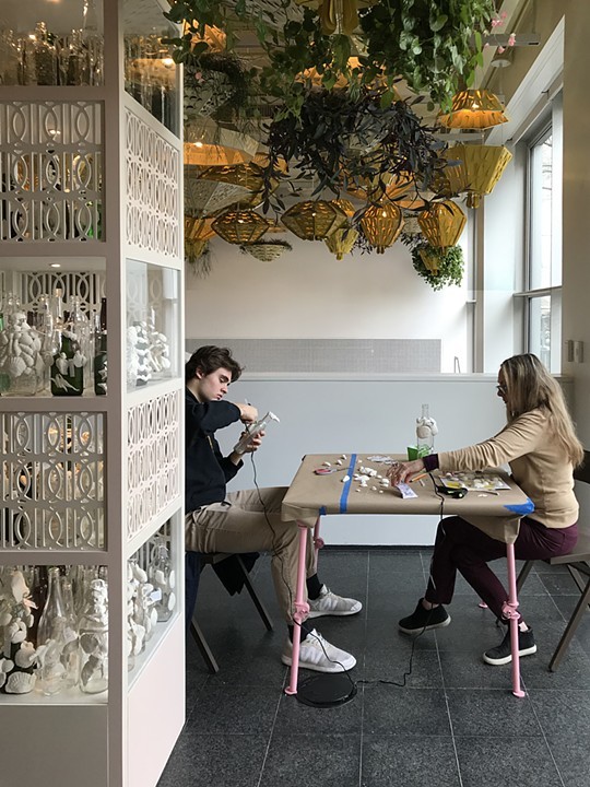 The image shows two people sitting at a table covered in shells and other miscellaneous craft supplies. They are decorating empty liquor bottles by gluing on shells. Next to the table is a shelf structure with empty liquor bottles as part of "Open 24 Hours." The activity pictured is part of the workshop series that accompanies Soto's installation at the MCA. Image courtesy of the artist.
