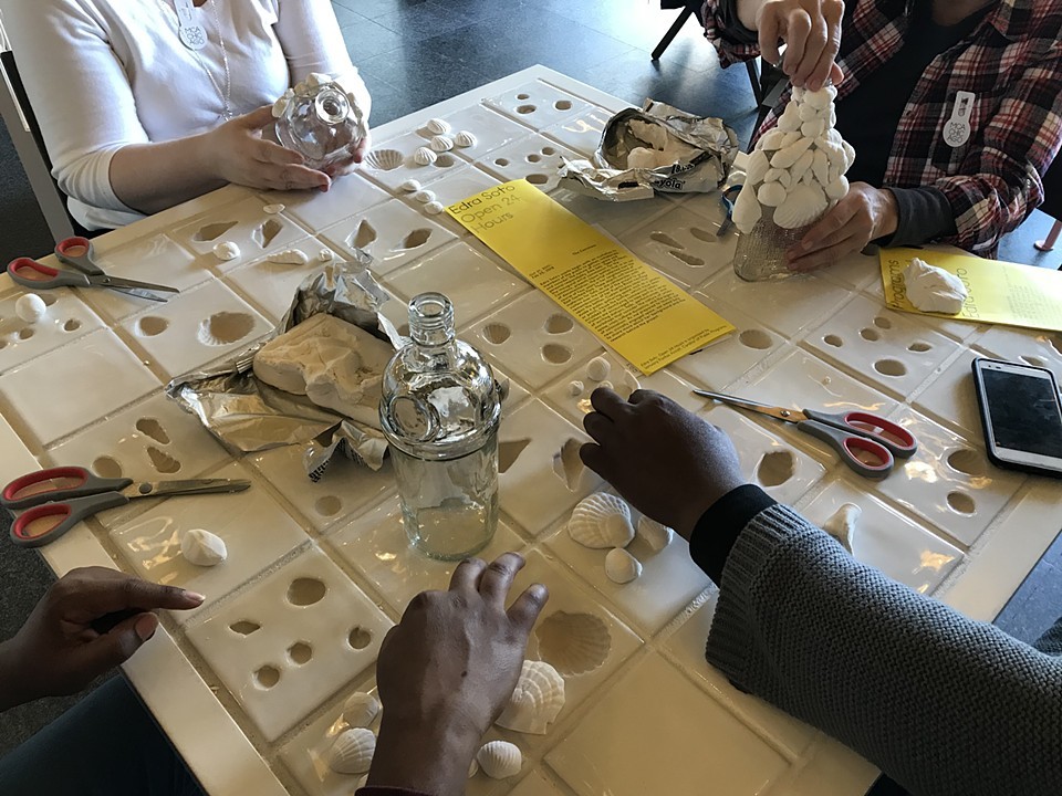 This image shows the hands of four people attaching shells to empty glass liquor bottles. The four people are sitting at a white table that has imprinted moulds in the shape of shells inlaid into its surface. On the table there are shells, empty liquor bottles, scissors, yellow pamphlets, and a cell phone. The activity pictured is part of the workshop series that accompanies Soto's installation at the MCA. Image courtesy of the artist.