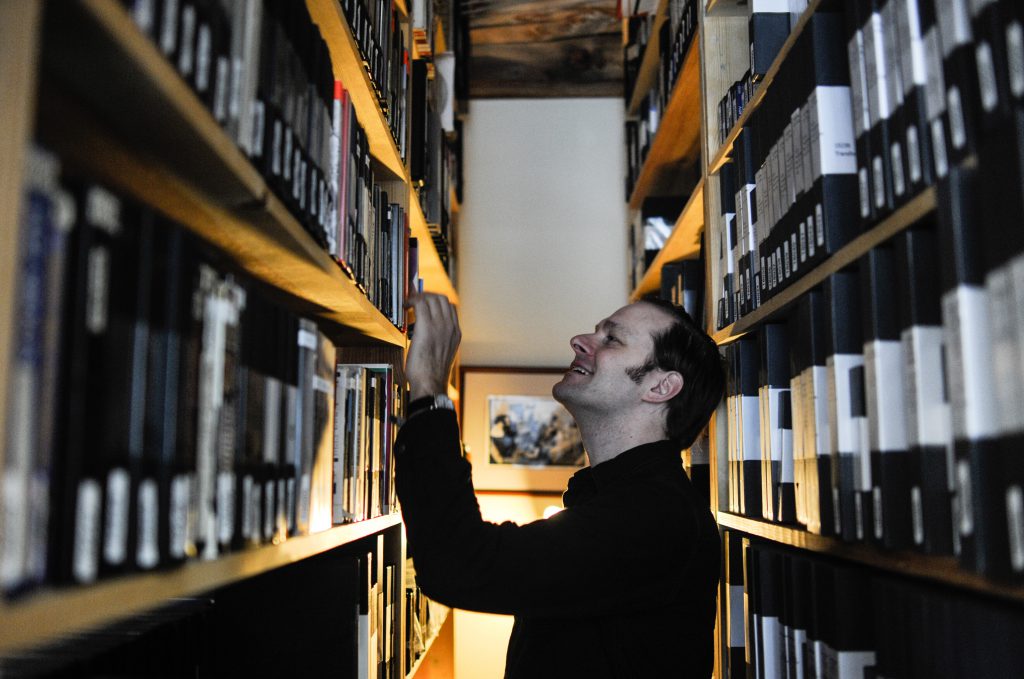 Photo by William Camargo. Dan Erdman at Media Burn is looking at the archival shelfs with one hand on a shelf. 