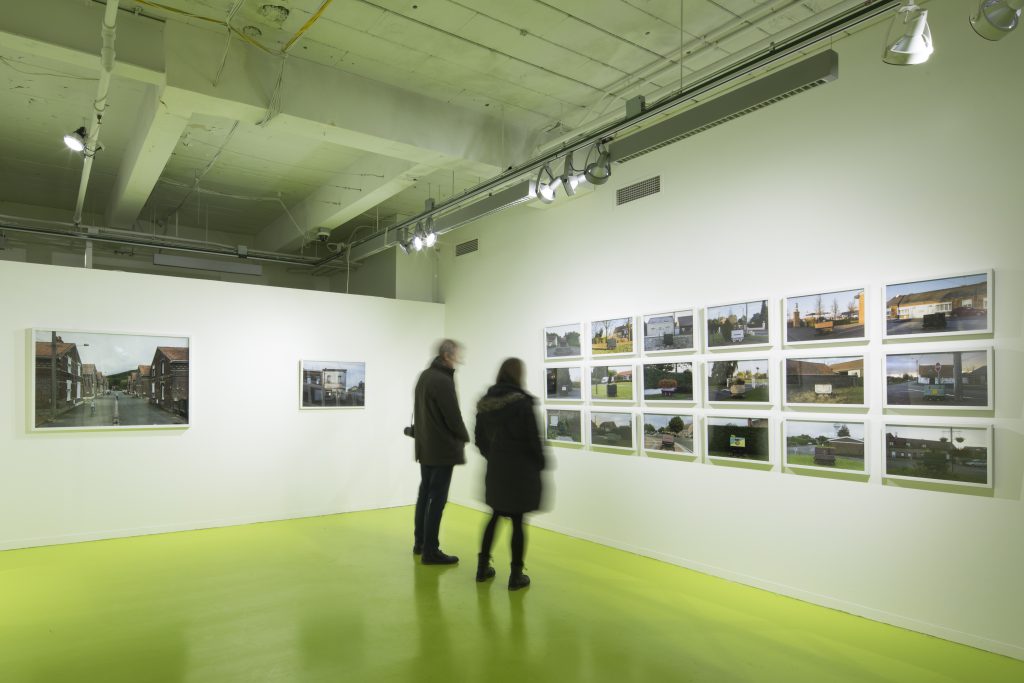 Two viewers take in David Schalliol's "Mining Basin (Hauts-de-France)". To their left are two isolated photographs of the town; on the wall in front of them is a series of images of the region's homes and distinctive coal carts, arranged in a grid that takes up most of the wall. Photo courtesy of Hyde Park Art Center.
