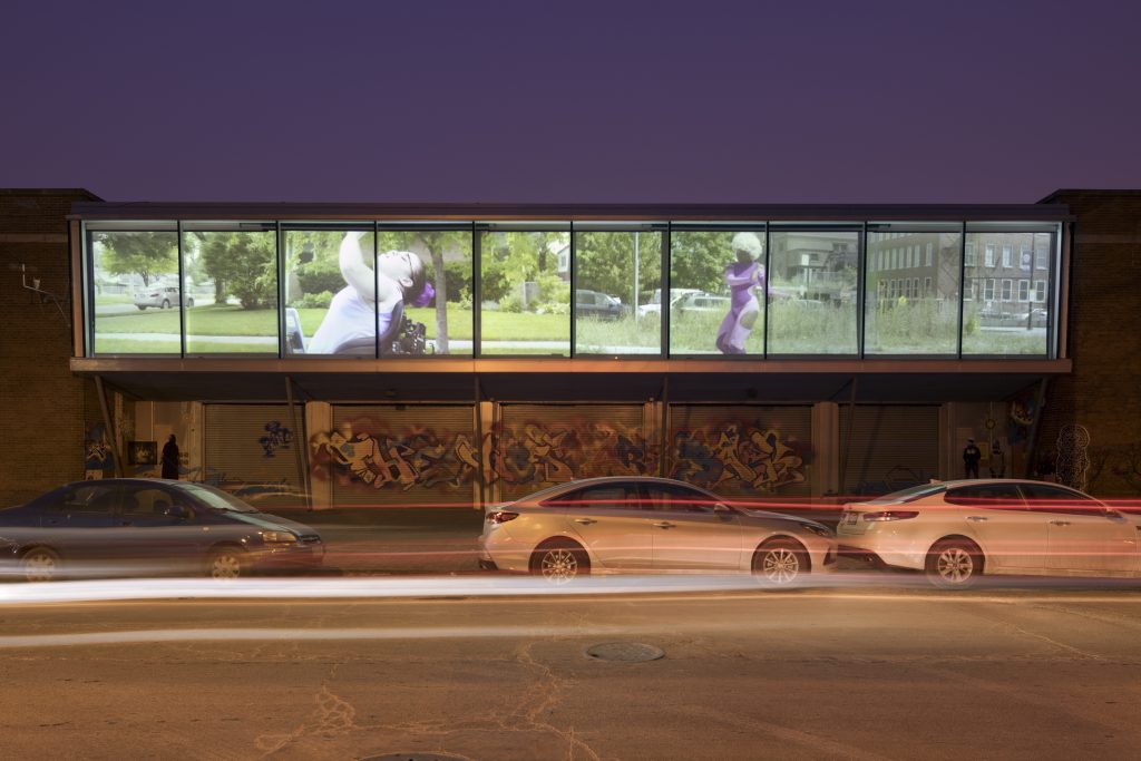 Justine Pluvinage's "Amazons" as seen from Cornell Ave outside the Hyde Park Art Center. A 10-panel display showing a grassy urban environment and a young woman is projected onto the building's facade. Photographer: 