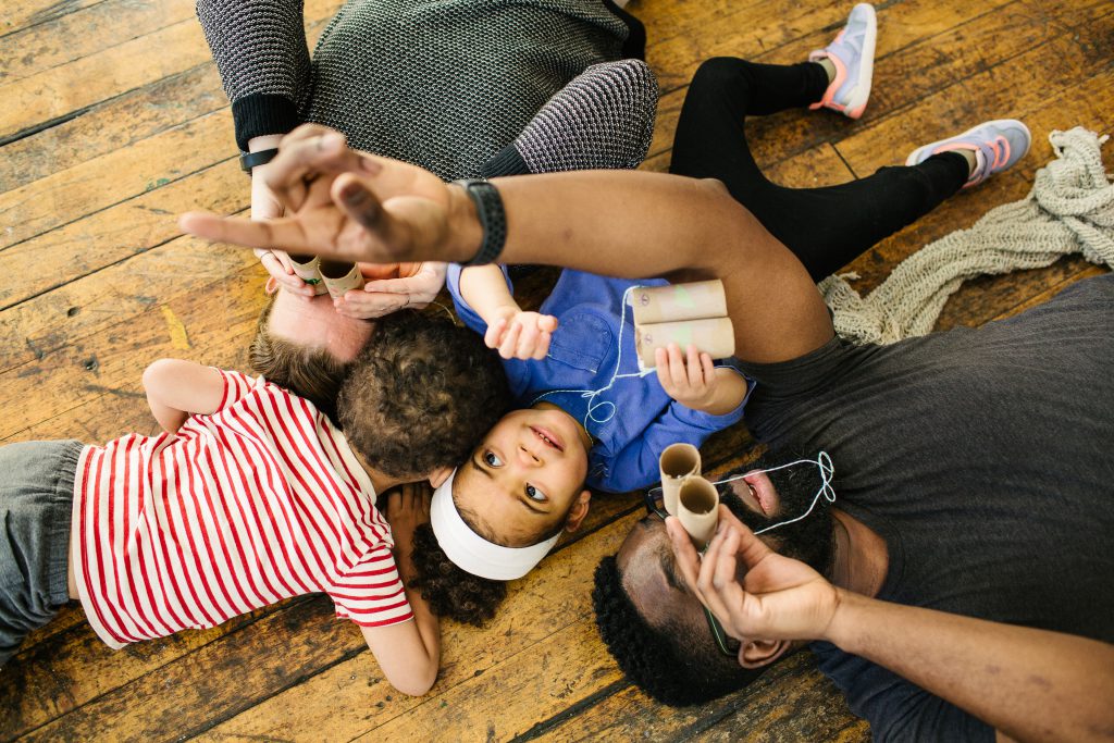 This photograph shows, from above, the artist, his wife Katie, and their twin toddlers lying on a wooden floor, with the adults and daughter holding cardboard tubes resembling binoculars and looking up. The artist’s left arm cuts across the image as he points toward the ceiling. His daughter’s eyes look where he’s pointing. Photo by Becca Heuer Photography.