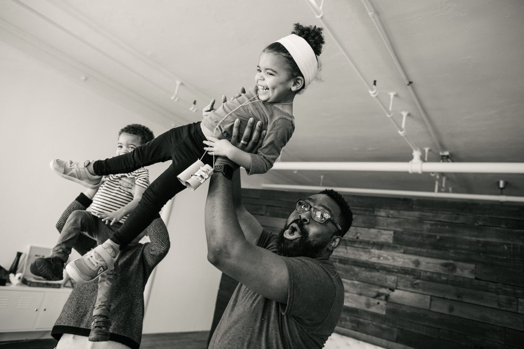 A black-and-white photograph of the artist, his wife, and their twin toddlers at play. They are in a spacious room with high ceilings. In the foreground, the artist lifts his daughter above his head and she laughs. In the background, his wife Katie lifts their son to her shoulder. Photo by Becca Heuer Photography.