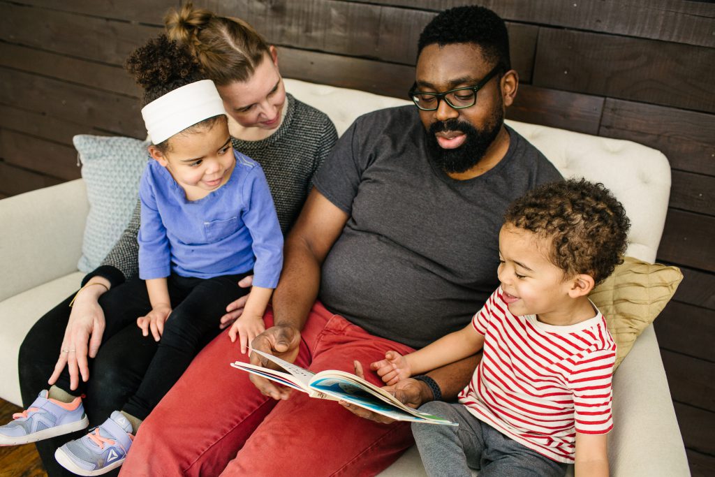 The artist, his wife Katie, and their toddler twins sit together on a large plush chair, in front of a wooden wall. Saleem holds a children’s book open in his lap and the others look on. His son points at the book and smiles. Photo by Becca Heuer Photography.