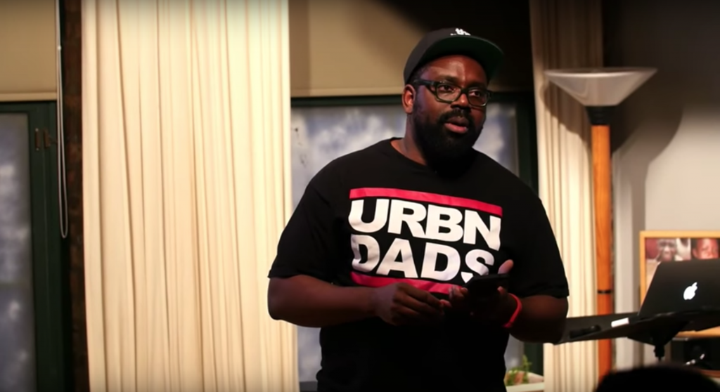 A photograph of the artist performing at Idea Potluck in 2016. He is wearing a black baseball cap, black glasses, and a black t-shirt that says “URBN DADS.” Behind him are curtains, a laptop, and photos. Courtesy of the artist.