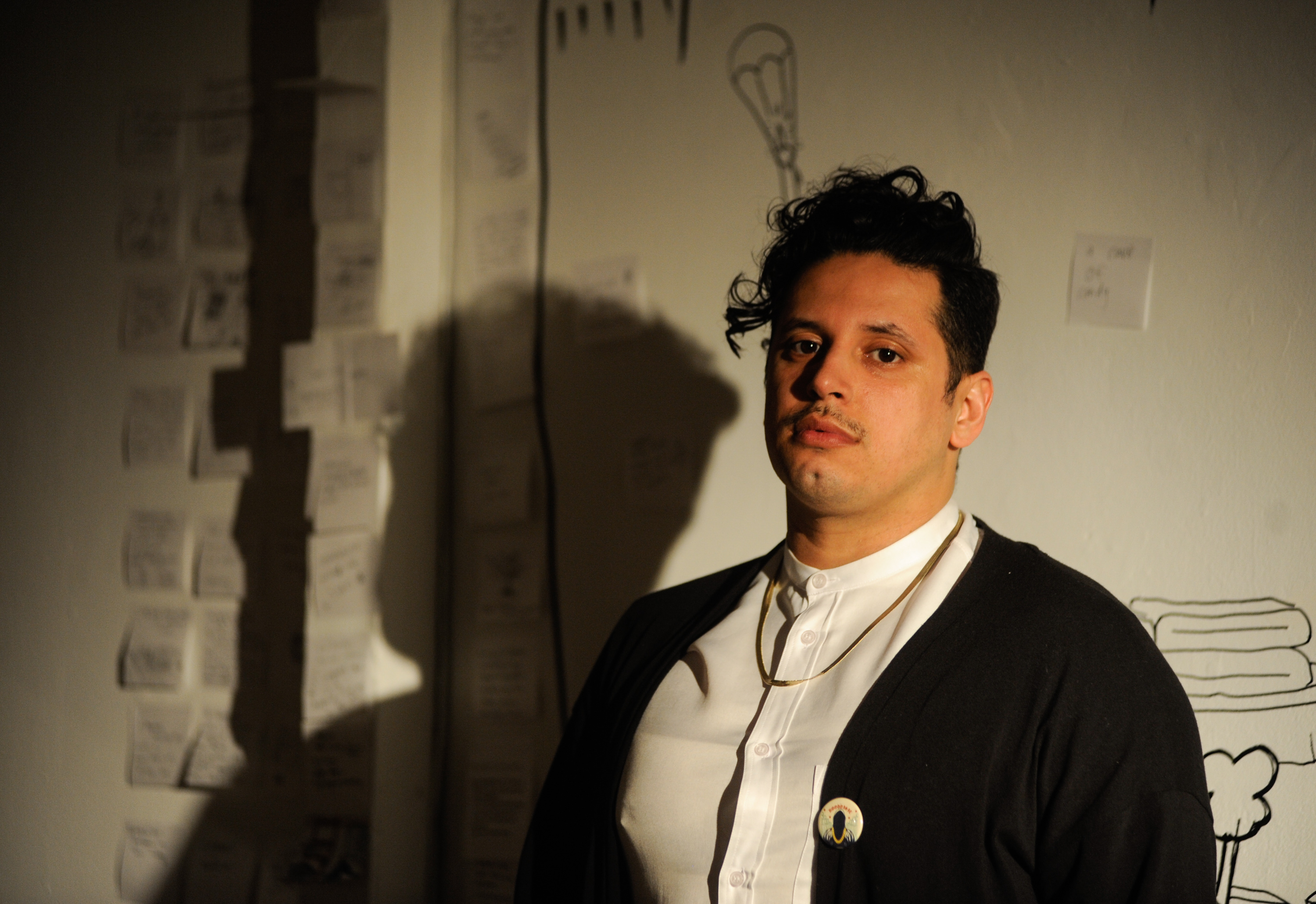 Ricardo Gamboa, host of the Hoodoisie, poses right before the start of February 3rd's episode at the Chicago Art Department. Gamboa wears a white button-up shirt, gold chain, and black sweater with a Hoodoisie pin. Their shadow is case on the white wall behind them. Photo by William Camargo