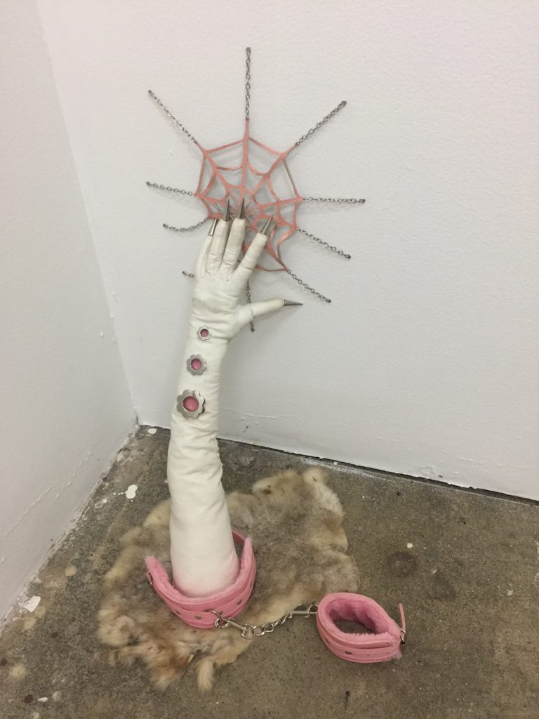 Courtesy of the artist. A white hand sculpture leans against the wall propping up a pink spider web. 