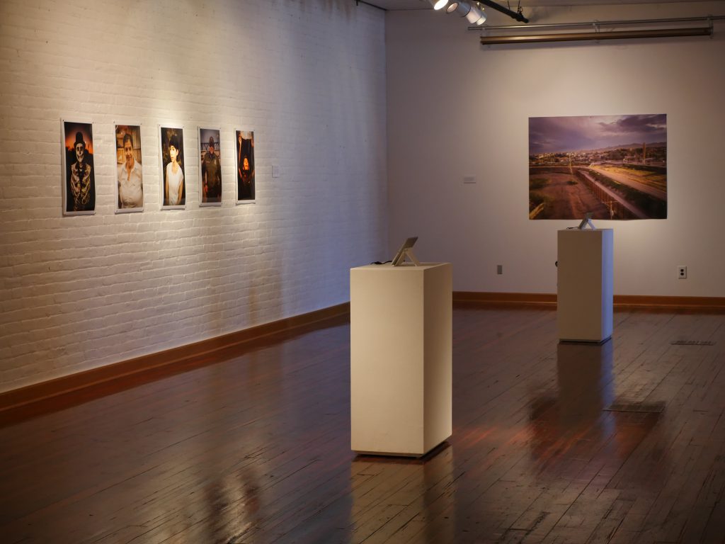 An installation shot of the exhibition shows portraits of El Paso and Juarez locals, a drone shot of landscape around the fence, and two iPads with videos playing. Image courtesy of the artists.