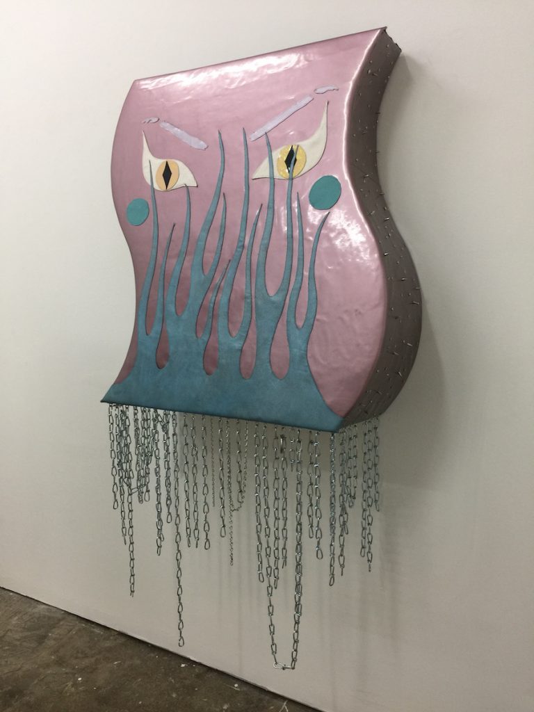 Courtesy of the artist. A large curved shape piece hangs on the wall with blue flames and yellow eyes. Chains hang from the bottom of the piece. 