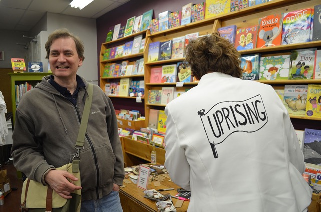 Image shows Nicole Garneau with a member of the audience. They stand next to a wall of children's books as well as the revolutionary women altar. She is wearing a white jacket with the word "UPRISING."