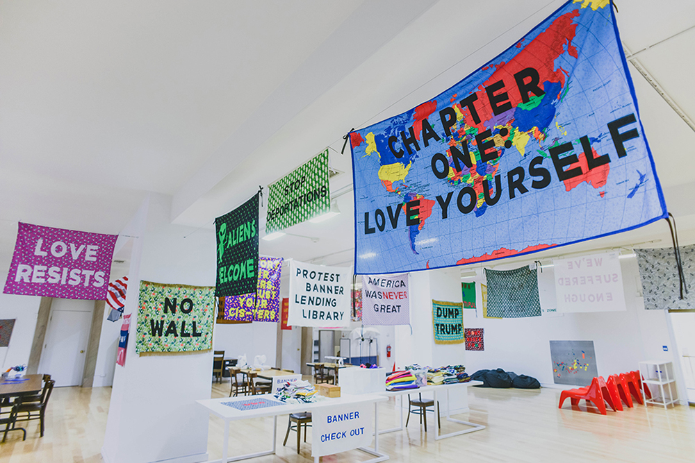 “Protest Banner Lending Library”, 2016–present. Installation of the “Protest Banner Lending Library” at the Chicago Cultural Center with protest banners hanging from the ceiling. Some of the most prominent banner read “CHAPTER ONE: LOVE YOURSELF,” “ALIENS WELCOME,” “NO WALL,” and “LOVE RESISTS.” Photo by eedahahm.