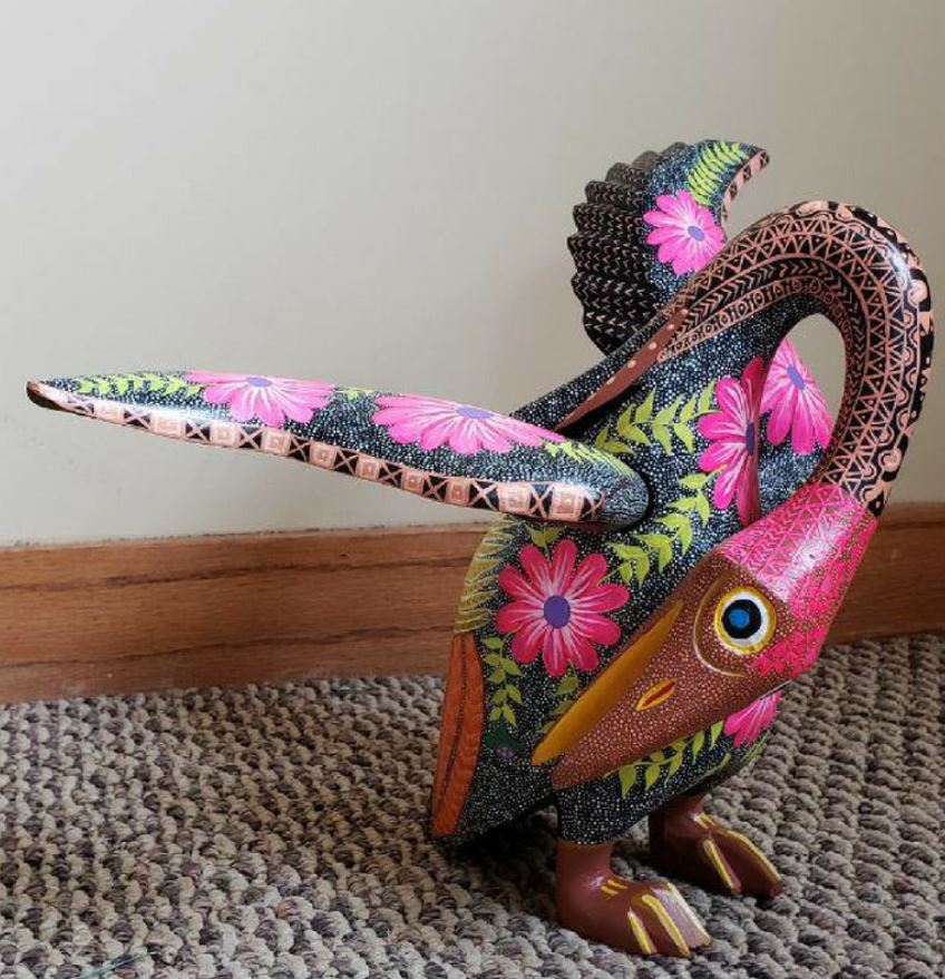 Alebrije sculpture by Carlos Orozco Ocuña. A carved bird curls its neck around its body and lifts its wings. The bird's head is bright pink and its body a stippled white and black with pink flowers. Image courtesy of the artist and Jennifer Smith of Cultura in Pilsen. 