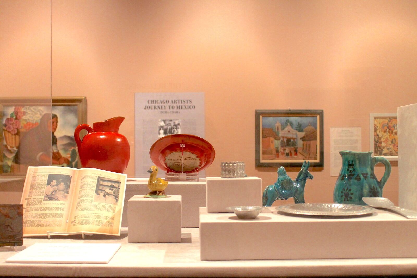Detail of a glass case from the exhibition holding silver dishware, ceramic jugs, and ceramic sculptures — one a yellow duck and another a green religious sculpture of Joseph walking behind Mary on a donkey. In the background there is a painting of an indigenous Mexican woman carrying flowers on her back in a basket, four photographs of Mexican artesanos working, and wall text that reads “Chicago Artists Journey to Mexico.” Image by Jennifer Patiño Cervantes.