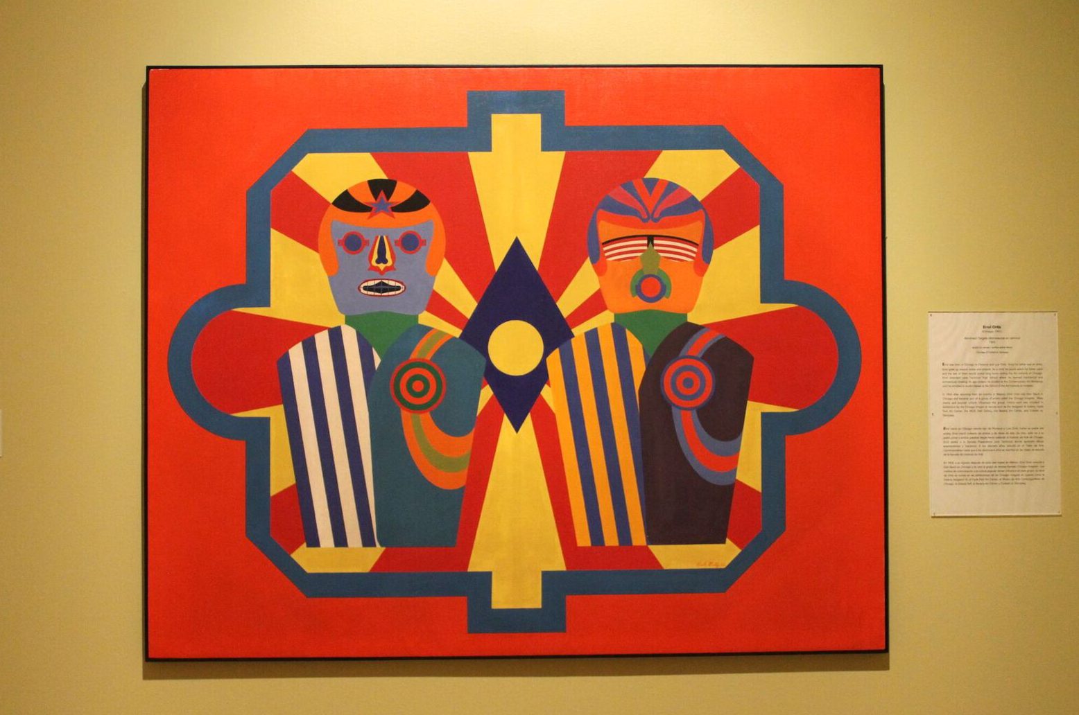 Astronaut Targets by Errol Ortiz. Red and yellow rays emanate from a blue and yellow diamond shape in the background of the painting. Two colorful figures wearing striped suits, and helmets with stylized features stand in the foreground side by side. Photo by Melissa Patiño Cervantes.