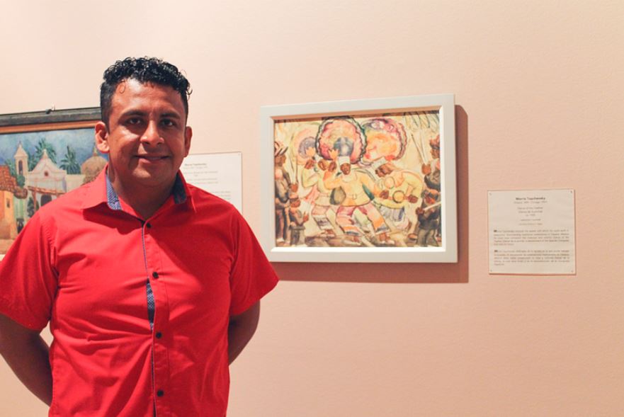 Artist Carlos Orozco Ocuña stands in front of the painting Dance of the Feather by Morris Topchevsky. In the painting, several figures dance or drum while wearing bright colored clothing and large round feathered headdresses. 