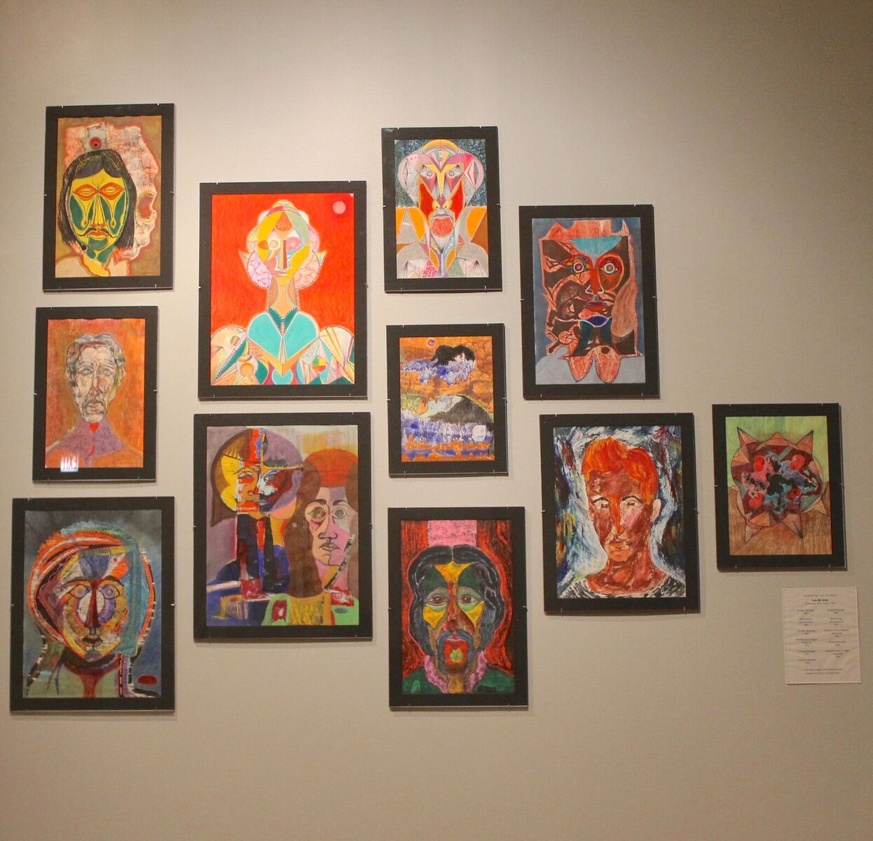 Detail of an exhibition wall showing portraits by Luis M. Ortiz. Eleven portraits are displayed. The portraits use flattened or angular planes for the faces and features, in alternating bright and earthy colors. In some places the paint on the portraits is rough and textured. Photo by Melissa Patiño Cervantes.