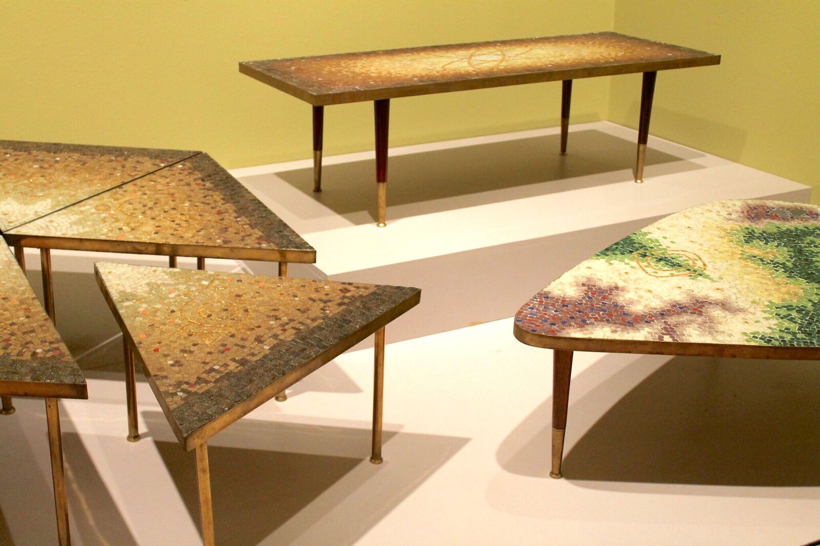 Mosaic tables by Genaro Álvarez . The table furthest back is long and has brown and gold tiles. Another table is a hexagon made up of smaller wedge shaped tables. And the table in the foreground is a triangular shape with rounded corners and red, purple, and green tiles. Image by Melissa Patiño Cervantes.
