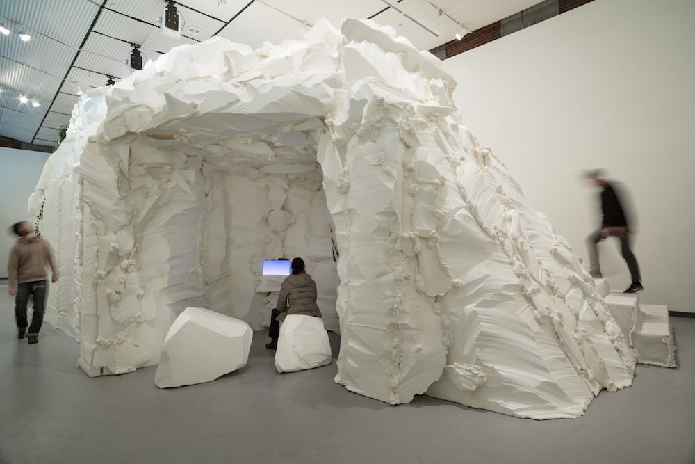 Installation view of "who cares for the sky" at Hyde Park Art Center, 2016. The image shows a large white mountain structure in the middle of a gallery space. Carved into the side is an opening with white rocks where a person is sitting and looking at an image on a screen. Photo courtesy of her website, sabinaott.com. 