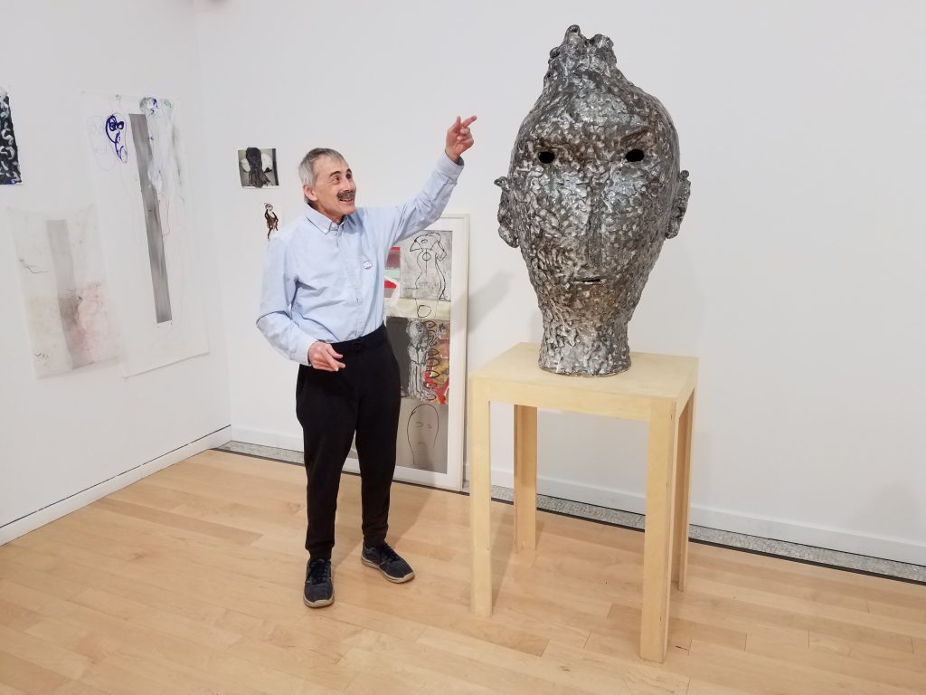 This photo shows one corner of the gallery. David smiles and points up toward the top of a tall sculpture, which sits on a narrow wood table. David, the sculpture, and the table are shown in full-view, close to straight-on, with abstract two-dimensional works visible behind them. The sculpture is silver and textured, with dark holes at the eyes and mouth. It has a slight nose, asymmetrical ears, and a tuft of hair at its top.