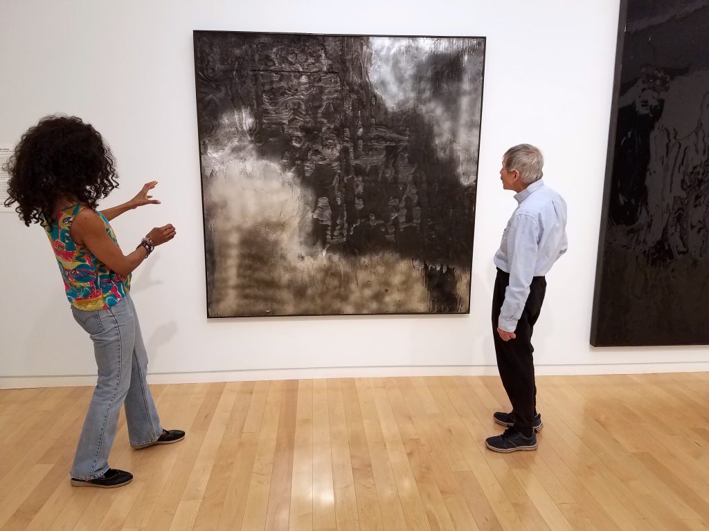 The poets stand in a gallery with an artwork between them—Lorraine with both hands up gesturing toward it and David looking at it, both with their backsides to the camera. The artwork is a large-scale, square piece, hanging on the wall. It is two-dimensional, with a textured black section stretching roughly diagonally from top-left to bottom-right corner, and with reflective silver sections in the top-right corner and along the bottom and in the bottom-left corner.