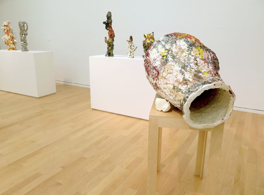 This photo shows part of the gallery. In the foreground on the right-hand side of the image, “Goya Dreaming” sits on a narrow wood table. The artwork is oblong in shape, with a colorful, textured surface, and a small whitish piece touching its side. From this angle, a large opening is visible. In the background are two broad pedestals (partially seen) displaying several sculptural objects.
