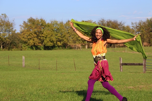 The poet strides across the frame—smiling, brightly-clothed, and with arms spread, a lime-green cloth suspended between her hands and floating behind her. She is outdoors in a grassy field, with a wire and wood fence in the near background and a line of trees farther back. The sky is light blue.