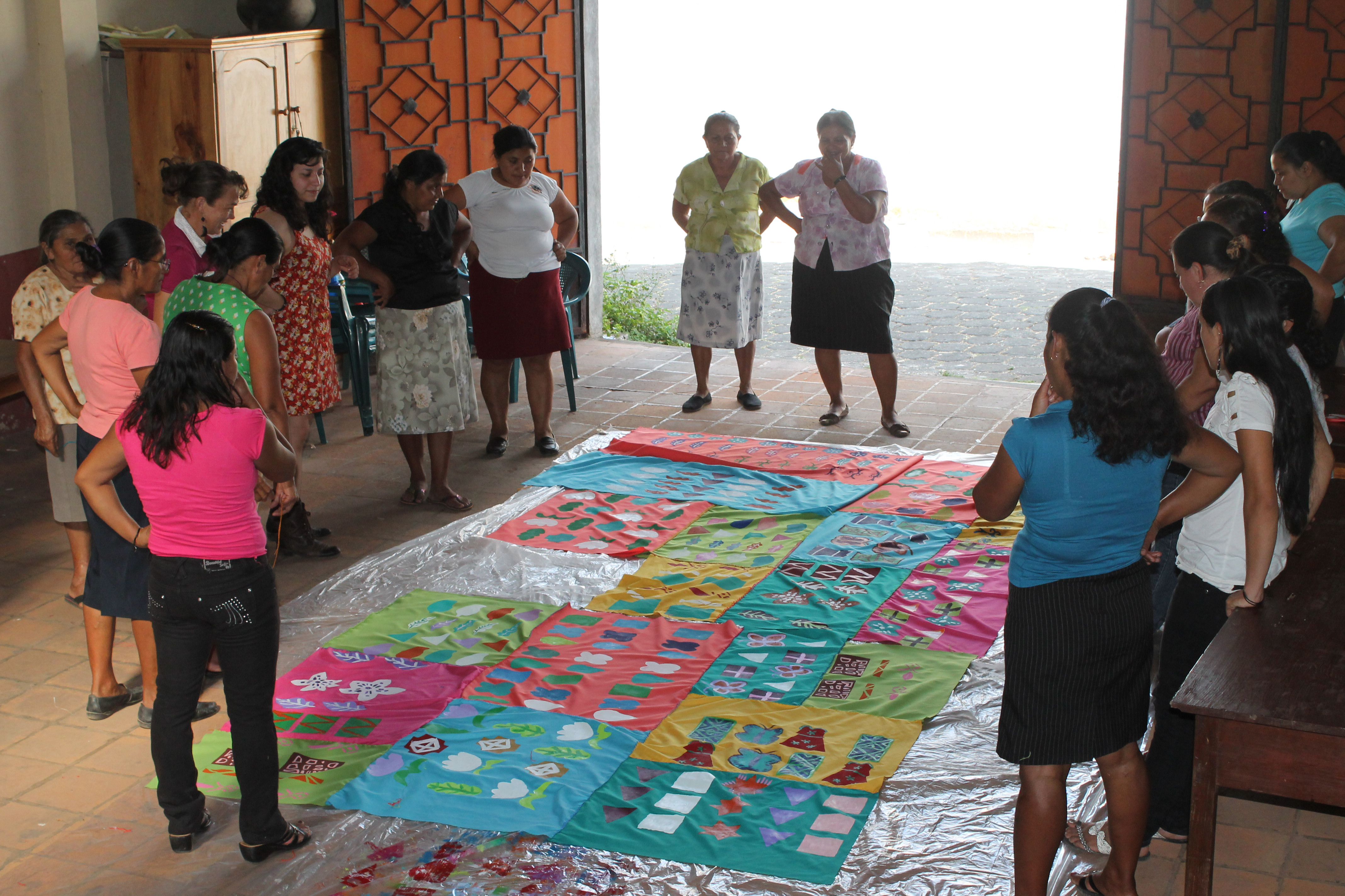 “Morazán Hilvanando Colores,” 2013. Fiber collaboration at Walls of Hope in Perquín, El Salvador. Victoria Martinez is standing with a group of women around a large textile on the floor. The textile is composed of different rectangular fabric pieces of varying vibrant colors that have a range of geometric patterns painted onto them. The piece was collaboratively made by the artists and these women, who have experience with war in El Salvador, from being nurses at war to having relatives to past survivors. 