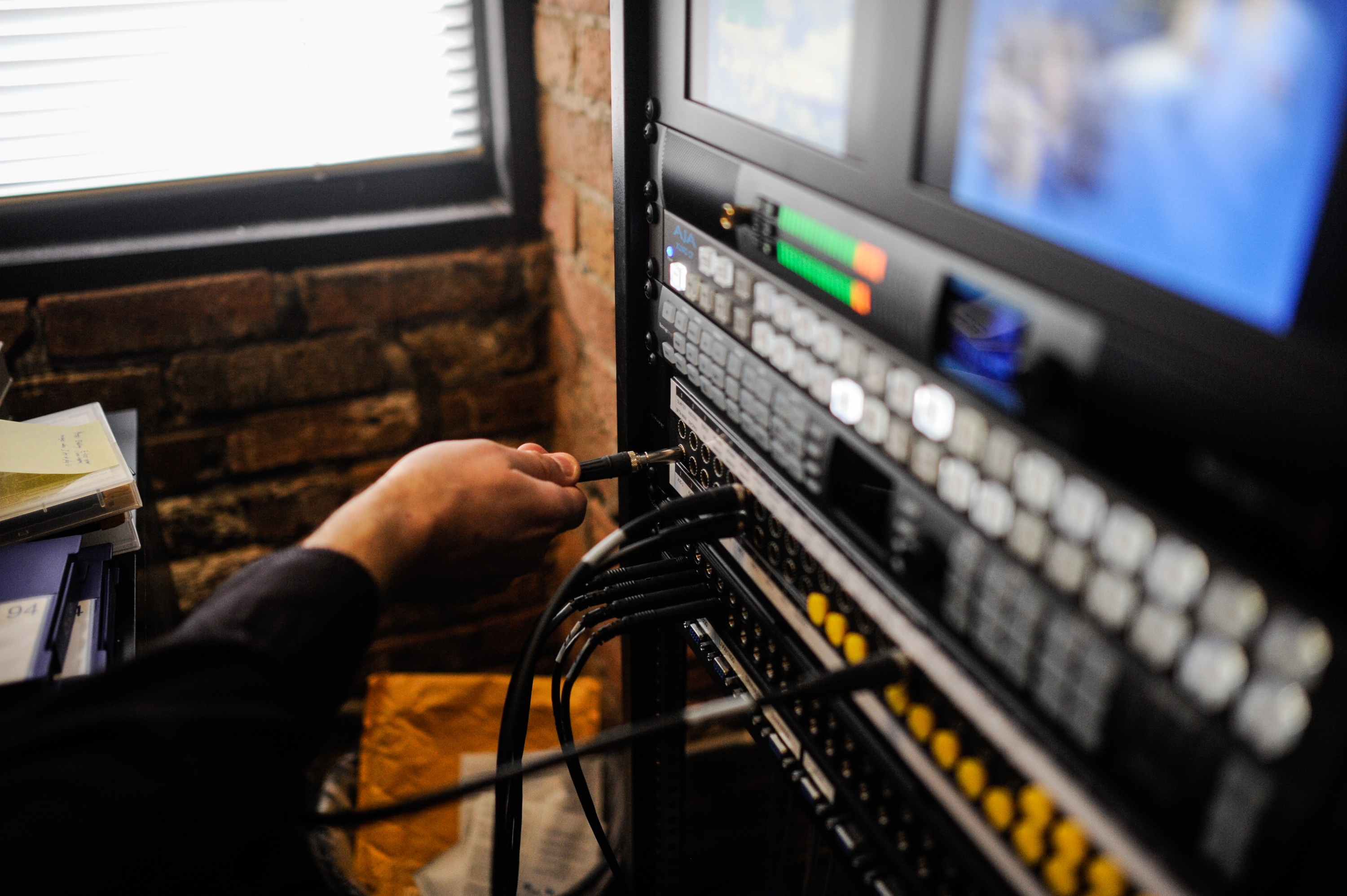 The image shows equipment at Media Burn archives. A hand plugs wire into the equipment below two screens. Photo by William Camargo.
