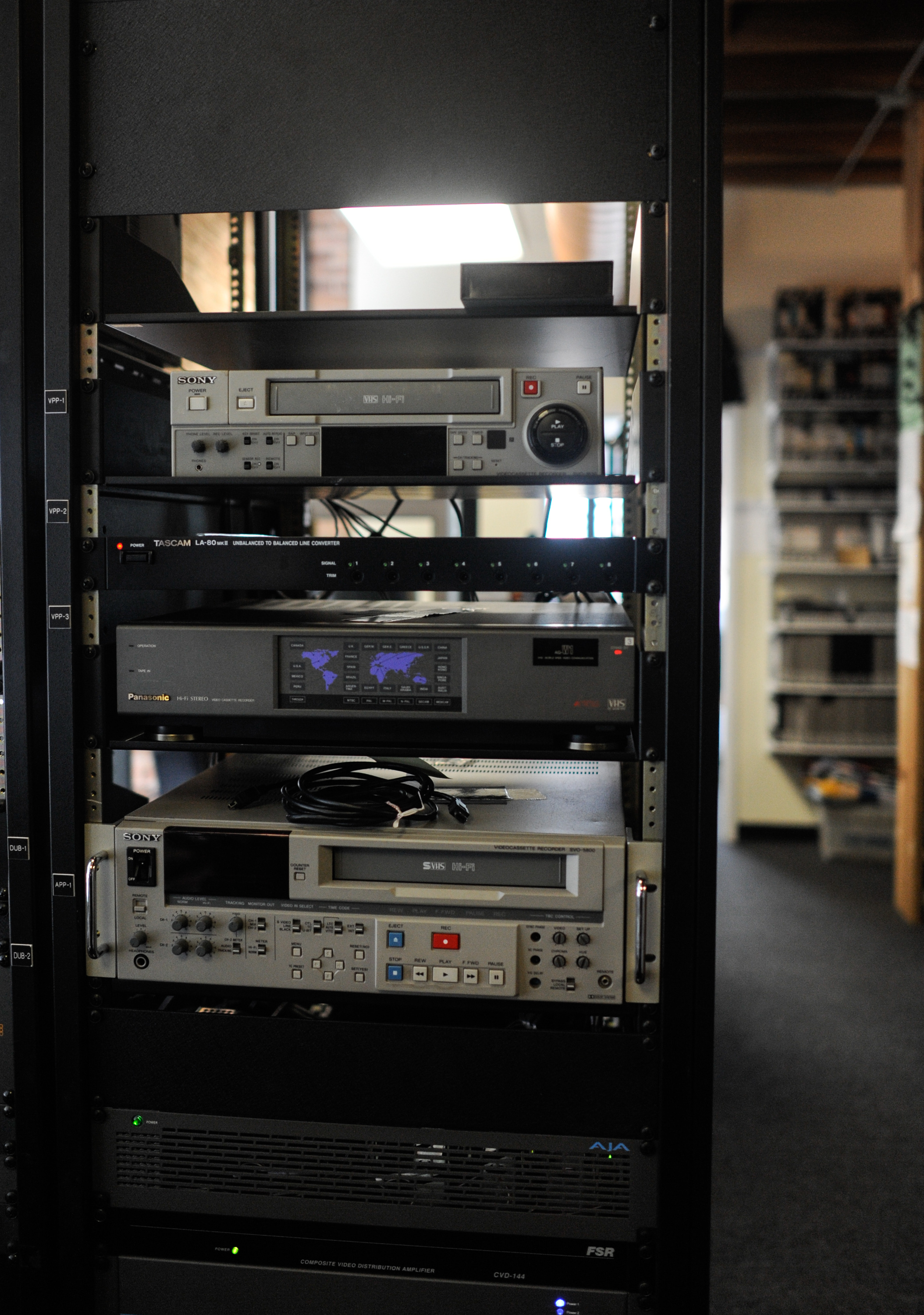 The image shows various equipment used for digitizing and playing different video formats. In the background, shelves hold video from Media Burn's collection. Photo by William Camargo.