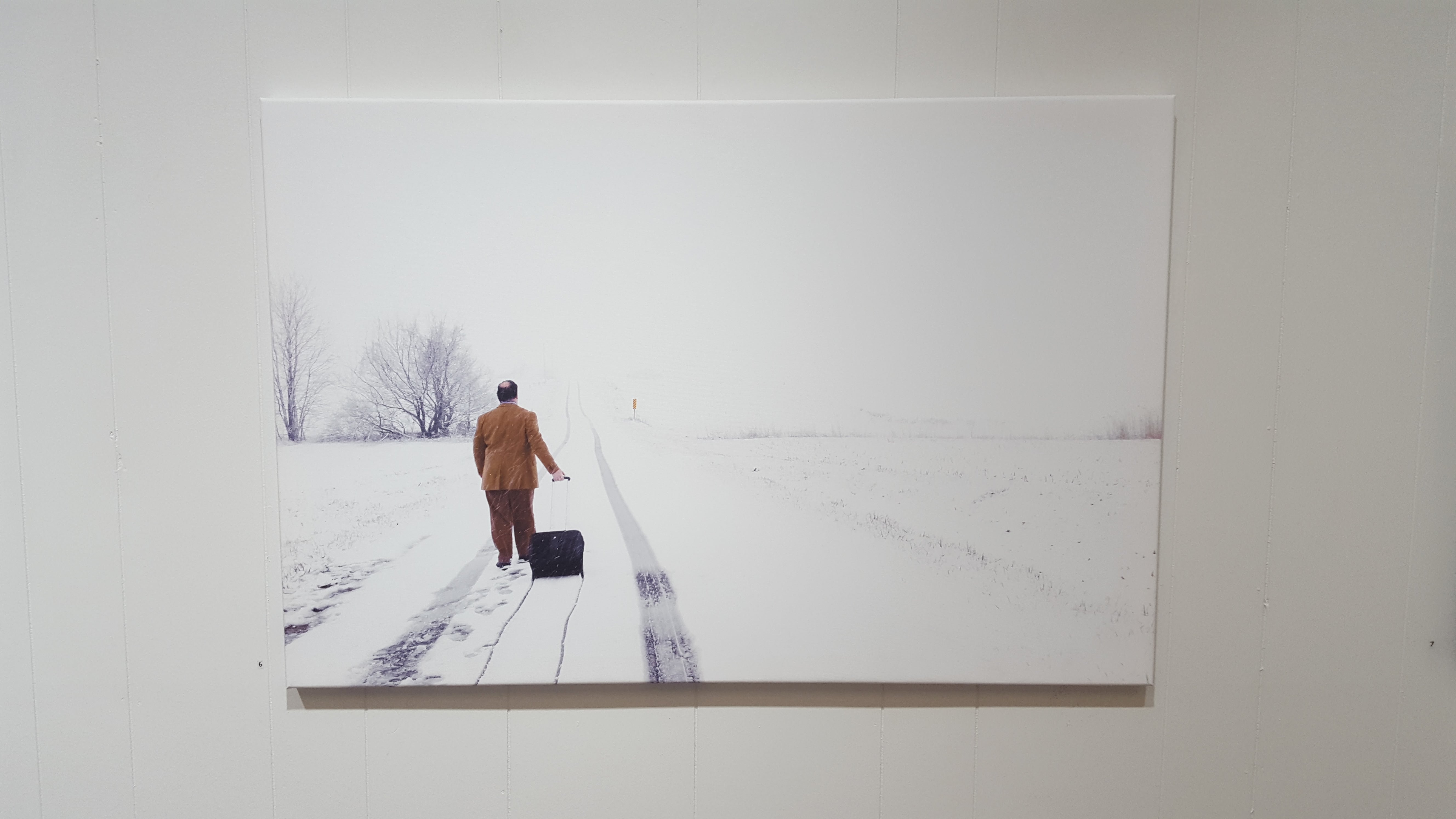 The artist is strolling down a snow covered road in business casual attire, pulling his suitcase behind him. Photo by Ted Diamond. Image courtesy of Edward Breitweiser.