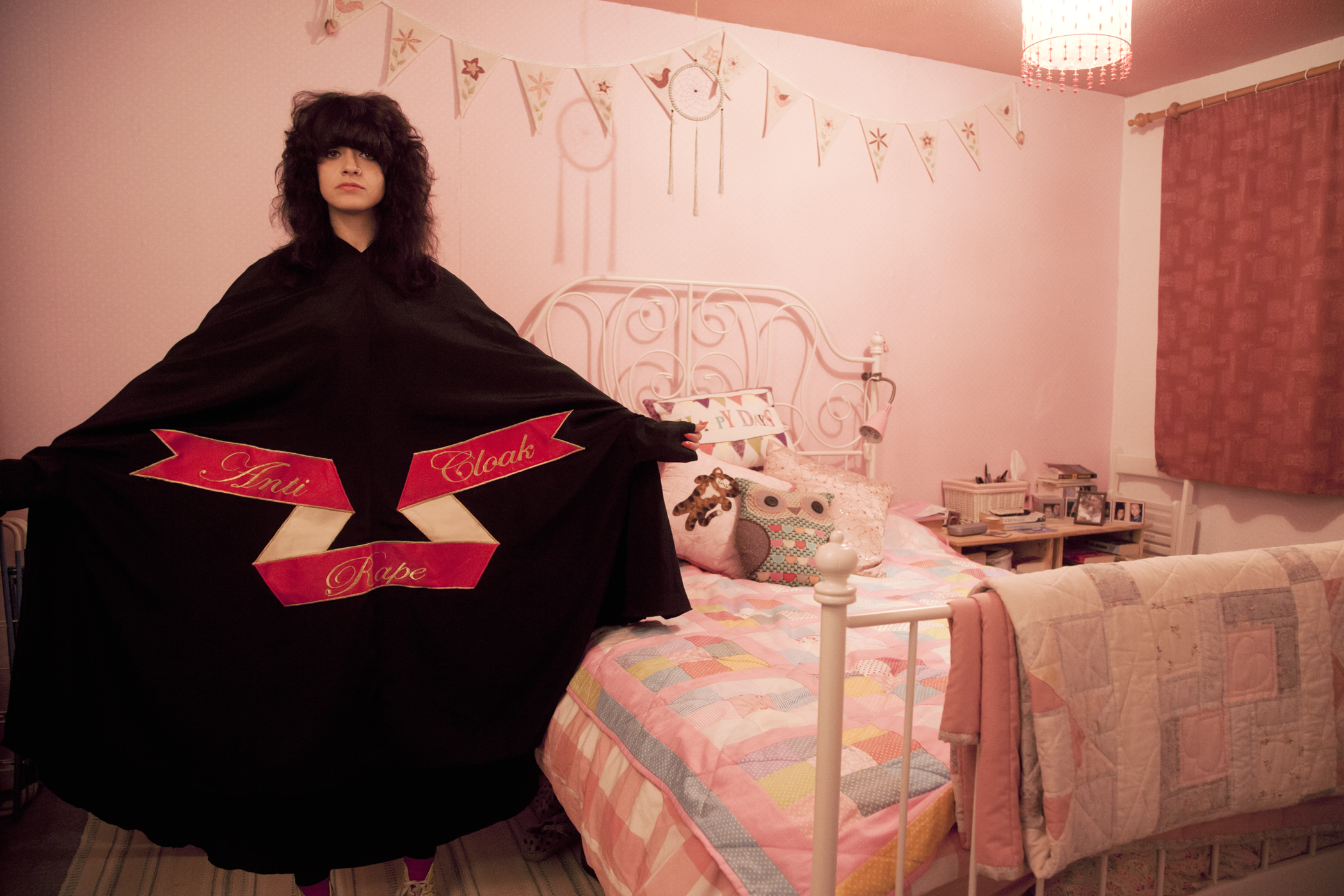 Sarah Maple, Bedroom Cloak, 2015. C-type matt photograph, 15.75 x 23.5 inches. Maple spreads her arms wide to show the text of her “Anti-Rape Cloak” as she stands in a pink bedroom. Photo courtesy of the Ukrainian Institute of Modern Art.