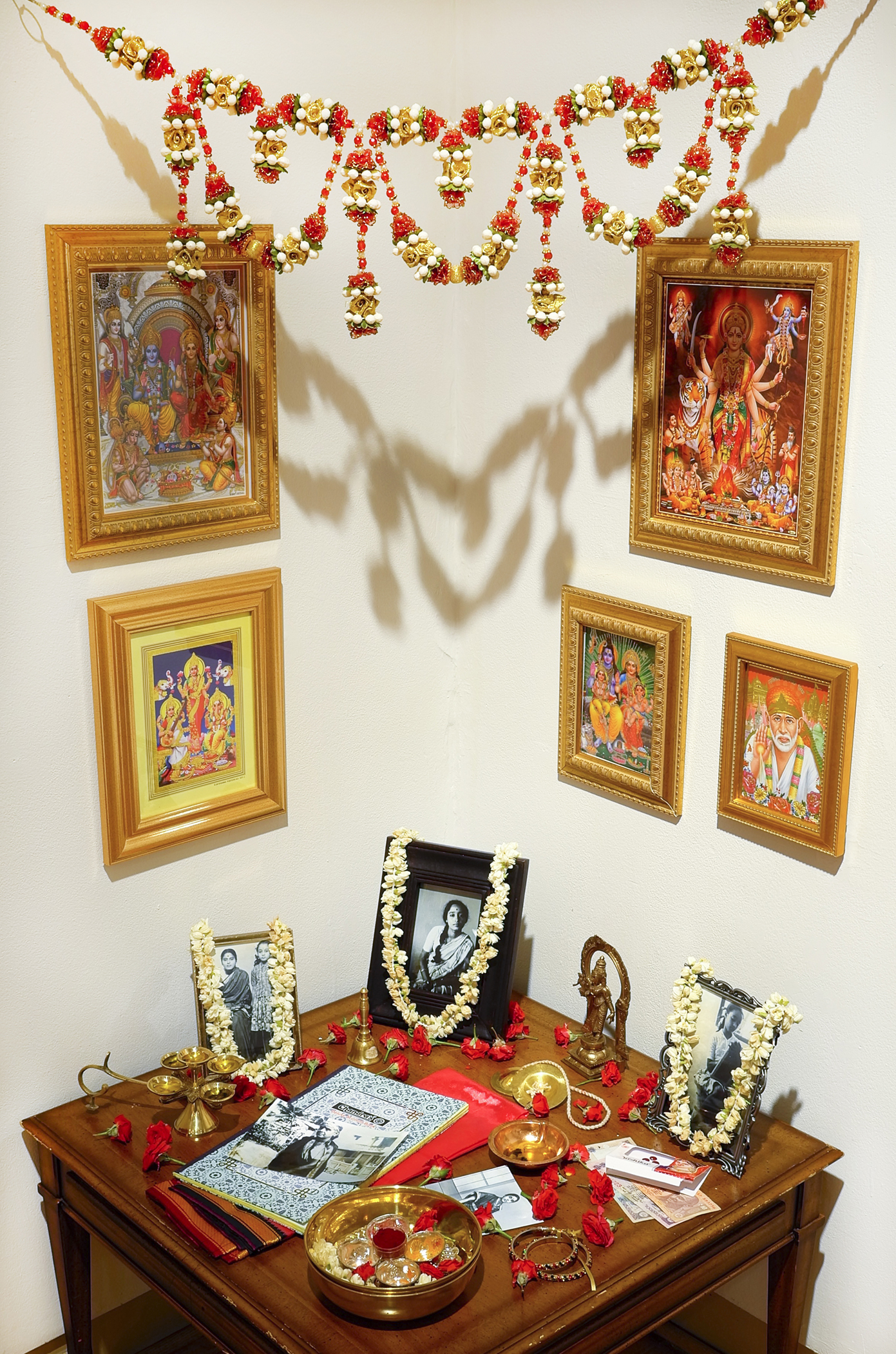 Lakshmi Ramgopal, Maalai, 2017. Table, written materials, framed photographs and prints, brass bowl, mixed metal statuette, hand bell, mp3 player, speakers. The devotional materials, including framed photos, dried flower chains, and a diary, rest on a small wooden table in a corner. Five colorful framed illustrations hang on the wall above the table along with a red, yellow, green, and white flower garland. Photo courtesy of the Ukrainian Institute of Modern Art.
