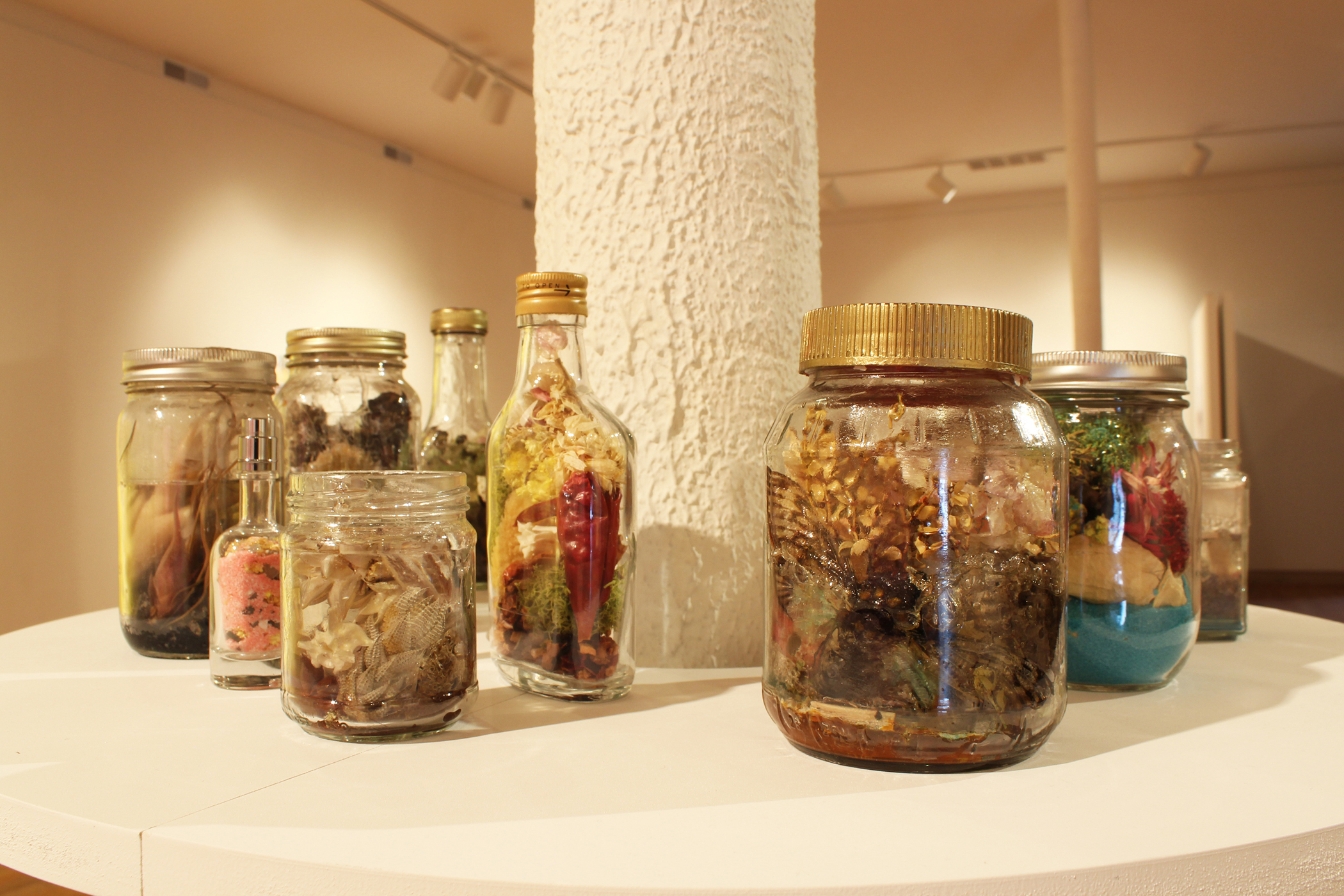 Rhonda Wheatley, Elixir Stills and Cure Bottles, 2017. Activate by gazing into jars and bottles. Gazing must be 100% voluntary. Healing intent and energy, incantations, secret herbs, sea horses, cicadas, snake bones, snakeskin sheddings, moss, dried flowers, sea shells, sand, wasp nests, succulent plants, various crystals (including amber, mica flakes, and pyrite), and more. Fourteen jars and bottles of assorted sizes rest on a round shelf encircling a column in the middle of the gallery. The clear vessels are filled with assorted organic materials, sand, and resin. Photo courtesy of the Ukrainian Institute of Modern Art.