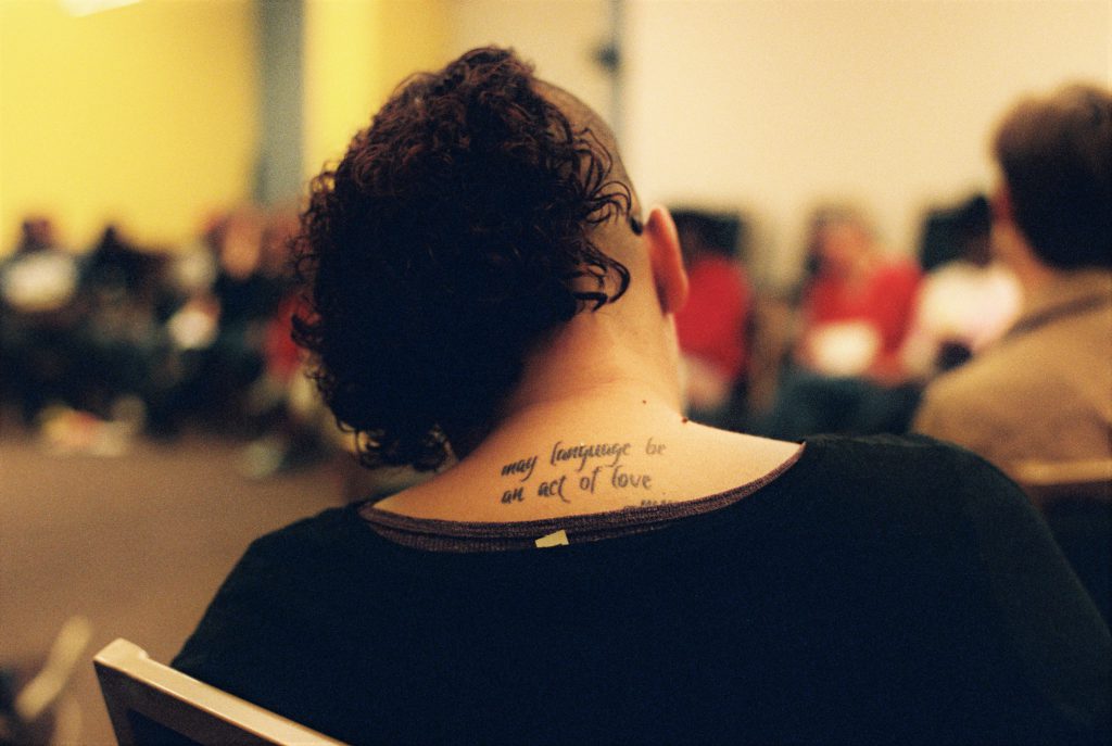 Image: A person with curly brown hair and a half-shaved head sits with back to the camera. A tattoo is visible on their back and reads, "May language be an act of love." Photo by Eric Roberts.