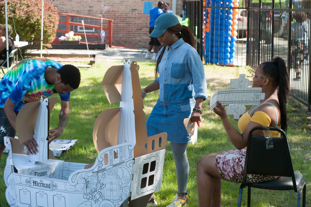 Image: Each Envisioning Justice community hub hosts several open houses throughout the residency. At the Brightstar Community Outreach open house in Bronzeville in May, a group builds a cardboard ship outdoors in the grass. Photo by Tony Smith.