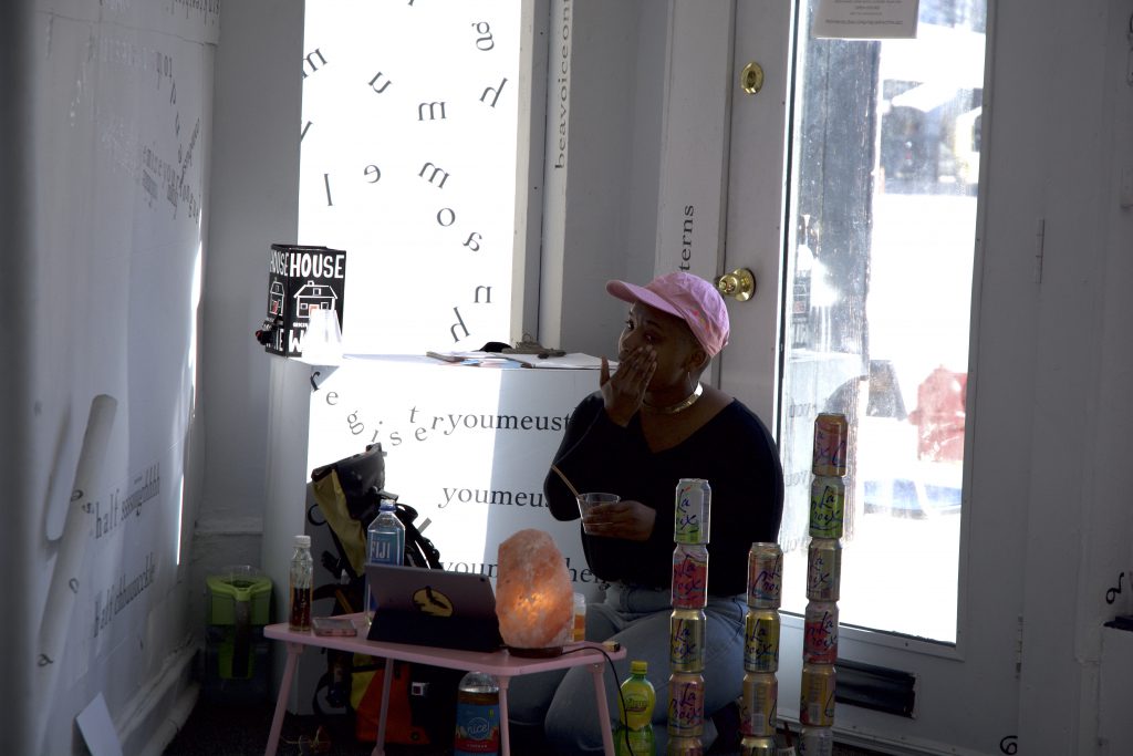 This is a still image of a performance. The performer sits inside the front door to the gallery, next to a wall covered in black vinyl letters and words. The performer wears a light pink baseball cap and sits behind a small, light pink table with a small, light pink, Himalayan salt lamp on it. Other objects near the performer include bottles of water and other liquids; towers of stacked, pastel LaCroix cans; and a cell phone and a tablet. The performer holds a small cup with one hand and spreads something on a cheek with the other.