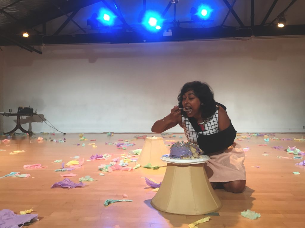 The image is a low, long shot of a performance space, with bright lights at the top of the frame shining toward the camera. In the foreground, Udita kneels on the floor, eating a bite of colorfully frosted cake off a plate that rests on a lampshade, also on the floor. Around and behind her in the room are shredded pieces of paper in pastel colors.