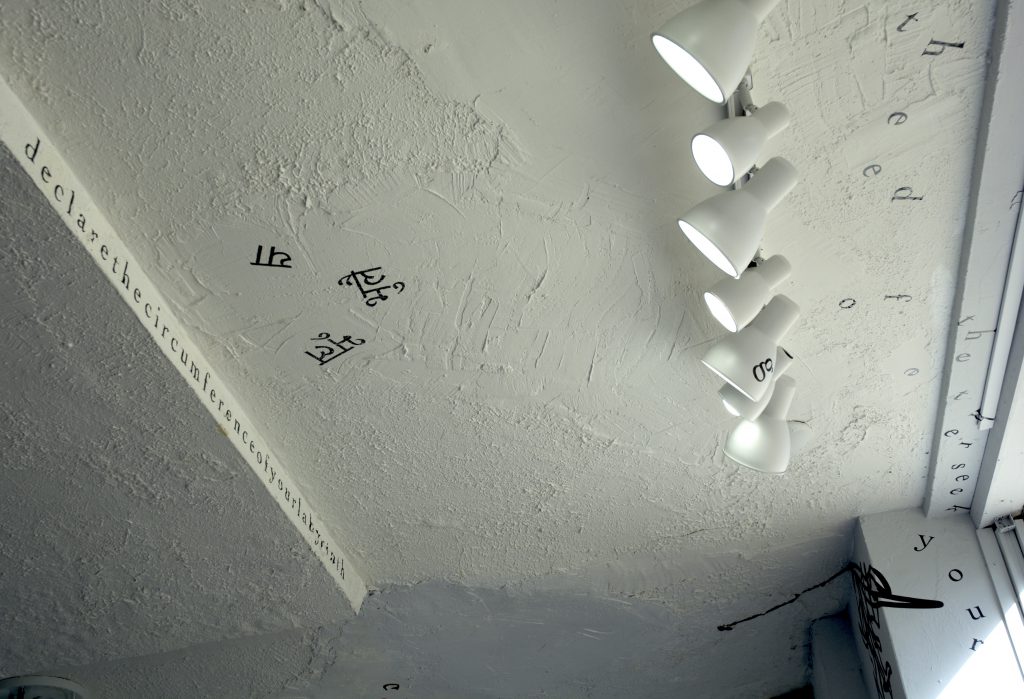 The photograph shows part of the gallery’s ceiling, a textured white surface with white track lights, and part of a window edge. Black vinyl letters are installed directly onto these surfaces and the walls, in the form of words and phrases in English and Hindi. Text appears in different sizes and spatial orientations (e.g., right-side up, upside-down, diagonal, vertical, and organic shapes), with some words/phrases expanded in space, condensed, or intersecting with other text. A gestural drawing—also made of black vinyl—is shown on the bottom right-hand side of the image.