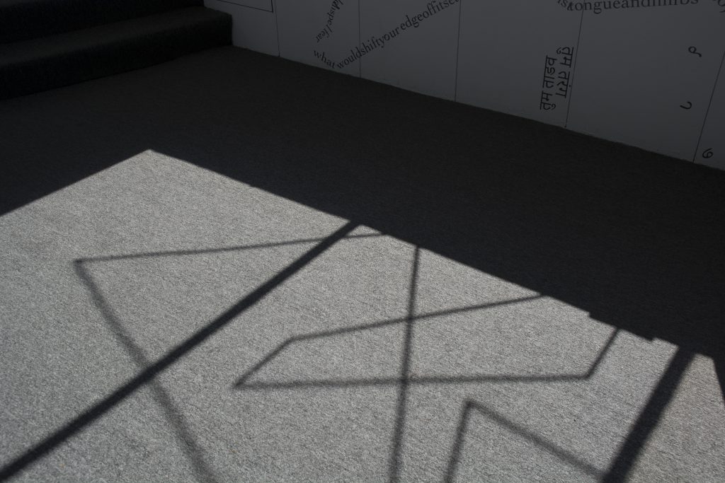 This photo shows part of the floor of the gallery, with sunlight from outside casting the shadow of the windowsill and a motif of intersecting vinyl triangular shapes onto a grey rug. Along the top of the image, black vinyl letters are partially visible on a white wall.